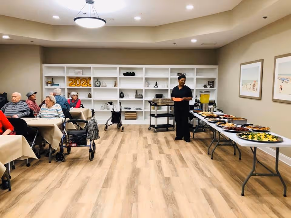 Seniors seated at tables in a community dining room with a buffet line and a staff member serving food.