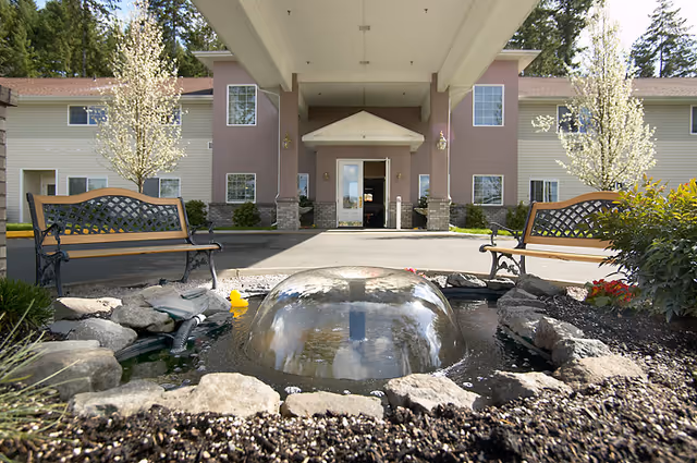 Entrance of a senior living facility with a covered driveway, two wooden benches on either side, a small water fountain with a dome-shaped water feature in the foreground, and blooming trees near the building.