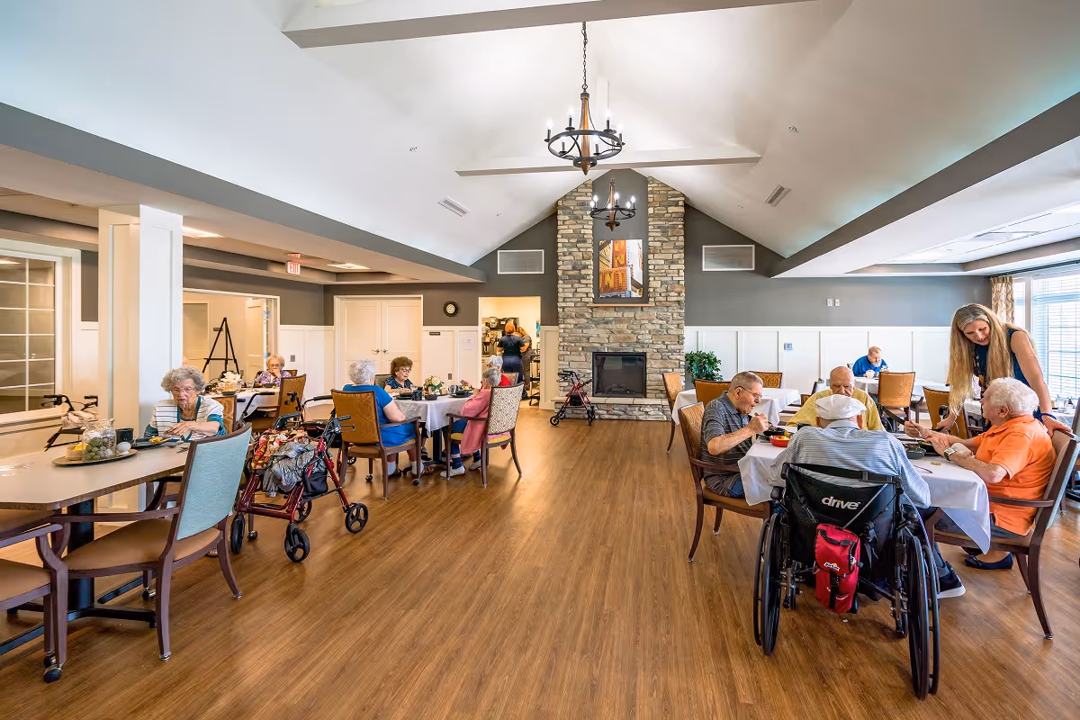 A spacious dining room in a senior living facility with several elderly residents seated at tables enjoying meals. The room features a high vaulted ceiling with two chandeliers, a stone fireplace in the center, and large windows allowing natural light. A caregiver is interacting with some residents at one table.