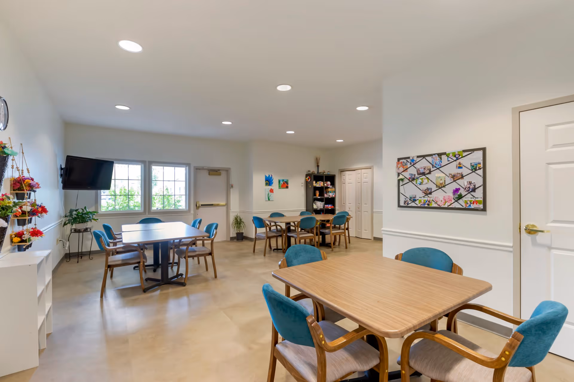 A bright and clean common room in a senior living facility with multiple wooden tables and teal cushioned chairs arranged around them. The room features white walls, a bulletin board with photos, a wall-mounted TV, and decorative plants. Large windows allow natural light to enter the space.
