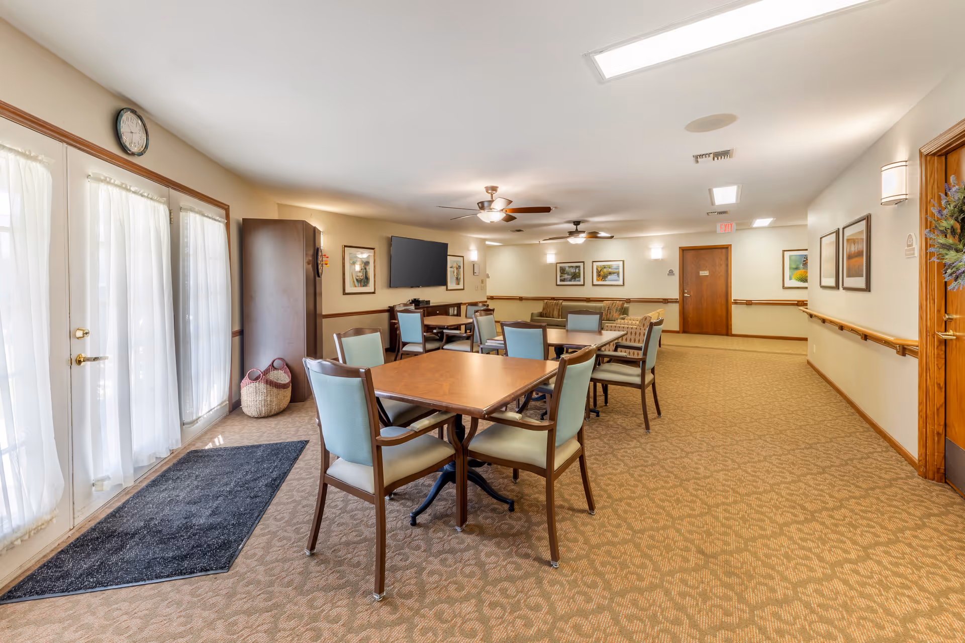 Well-lit communal dining/activity room with tables and chairs, a wall-mounted TV, and double glass doors.