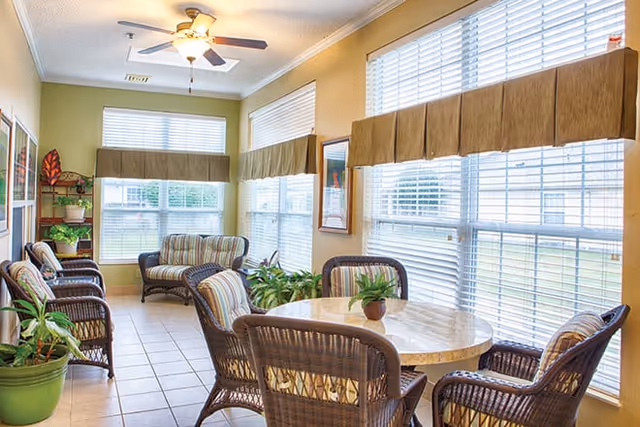 A bright and cozy common area with large windows covered by brown valances, allowing natural light to fill the room. The space features a round table with four wicker chairs around it, several potted plants, and additional wicker seating along the walls. The walls are painted a warm yellow, and a ceiling fan with lights is mounted above.