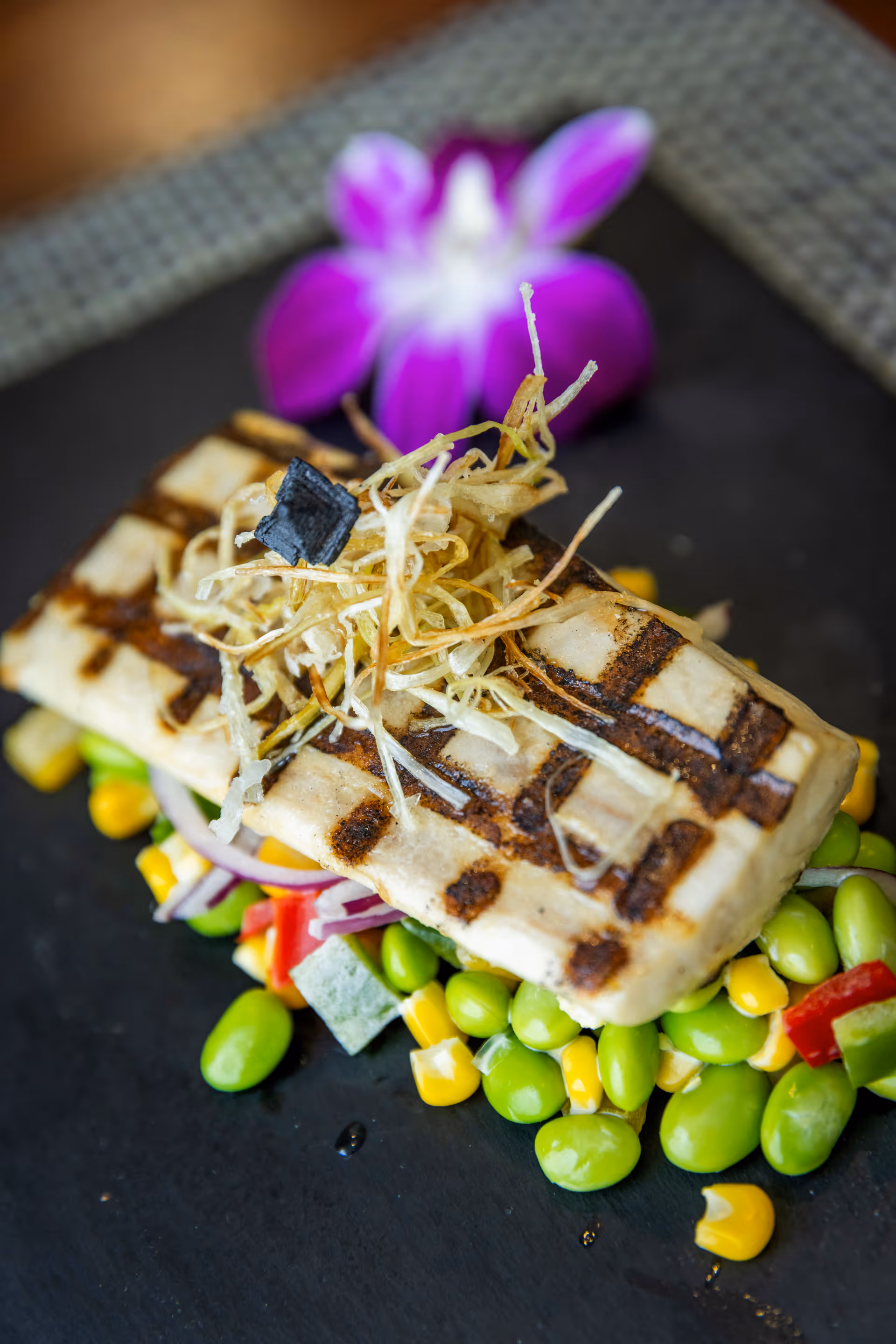 Close-up of a grilled tofu dish served on a bed of edamame, corn, and diced vegetables, garnished with crispy fried strips and a purple orchid flower in the background.