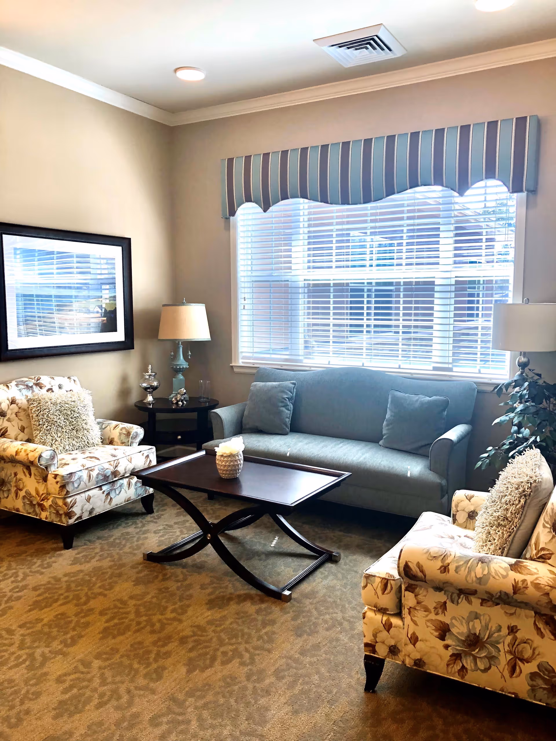 A cozy living room with a blue sofa, two floral armchairs with decorative pillows, a dark wooden coffee table with a small decorative item on it, a side table with a lamp and decorative pieces, a large window with white blinds and a blue striped valance, beige walls, and patterned carpet.
