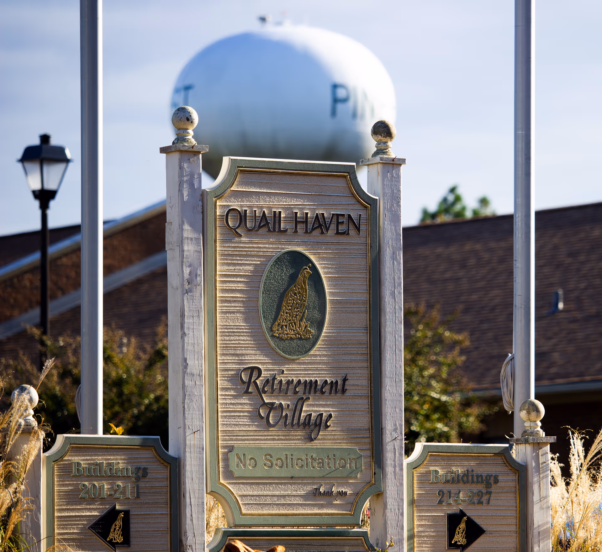 Wooden entrance sign reading "Quail Haven Retirement Village" with decorative posts and directional panels, a water tower and building roof visible behind.