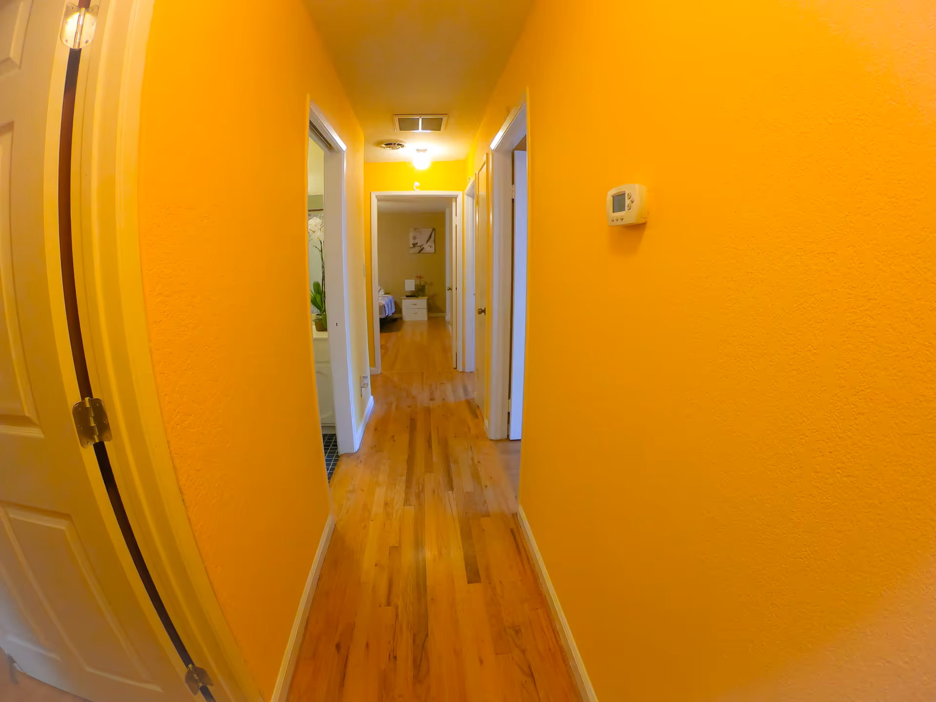 A narrow hallway with bright yellow walls and wooden flooring. There are several doorways along the hallway, with one room visible at the end containing a bed and nightstand. A thermostat is mounted on the right wall.