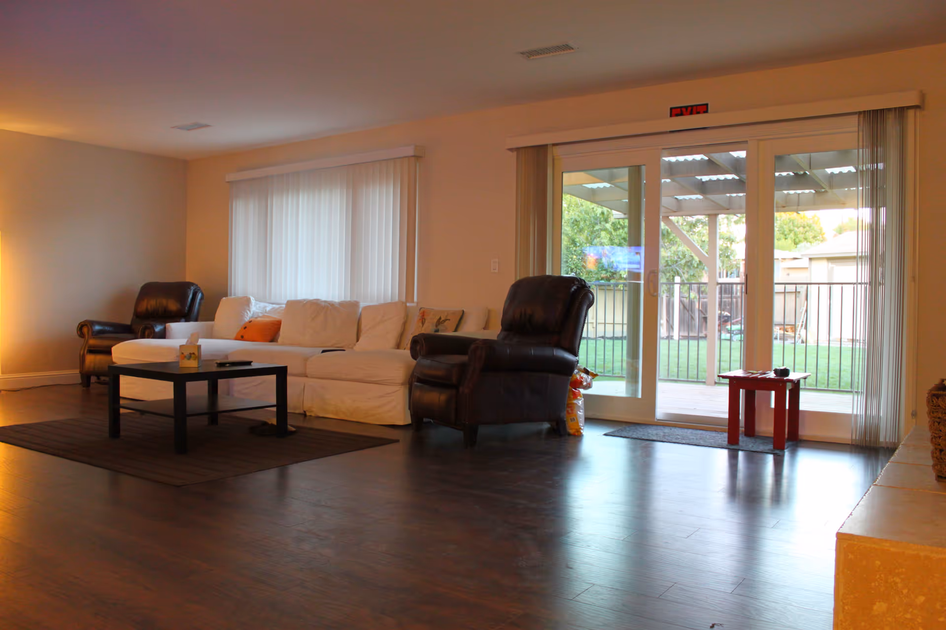 A cozy living room with a white sofa, two dark leather armchairs, a black coffee table on a brown rug, and large sliding glass doors leading to a covered patio and green outdoor area.