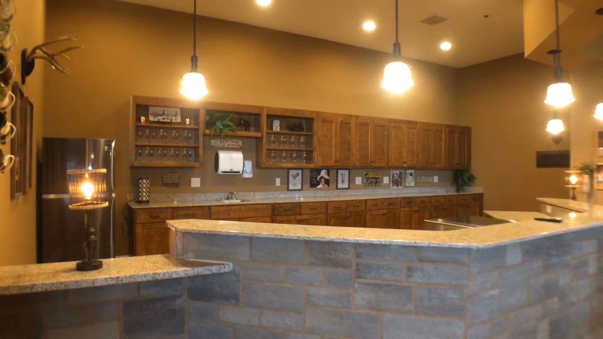Interior view of a kitchen area with wooden cabinets, granite countertops, and pendant lights hanging from the ceiling. There is a stainless steel refrigerator on the left, a paper towel dispenser mounted on the wall, and various decorative items and framed pictures on the wall above the counter.