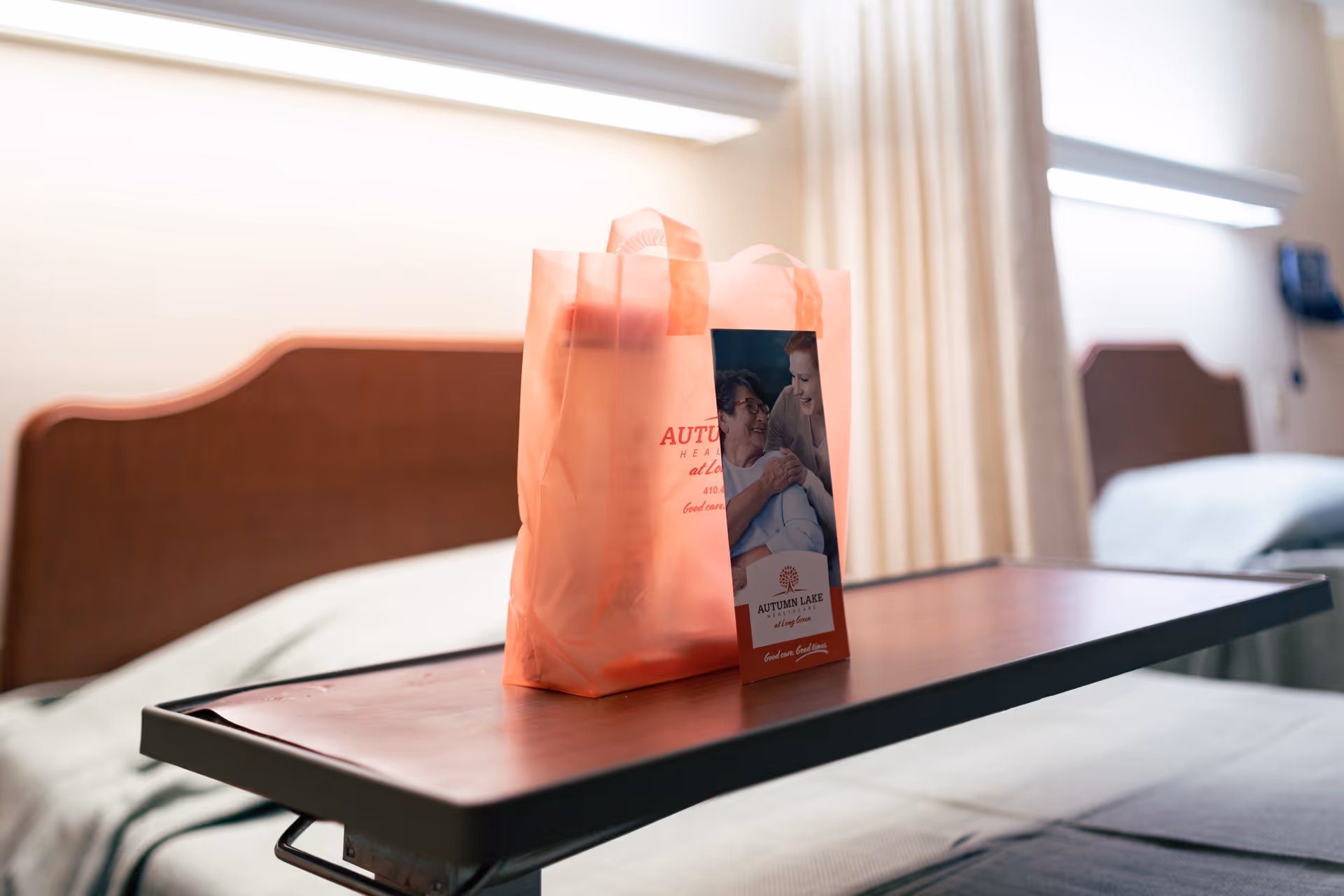 A hospital or nursing home room with two beds, a wooden headboard, and a bedside table with a pink translucent bag and a brochure from Autumn Lake Healthcare at Long Green placed on it. The room has beige curtains and soft lighting.