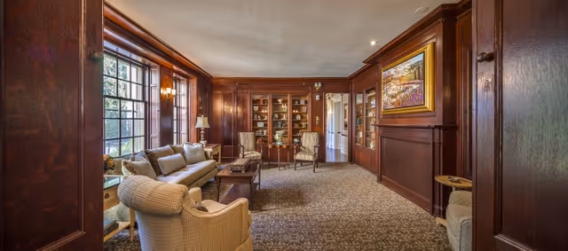 Warm wood-paneled sitting room with upholstered sofas and chairs, built-in bookcases, and a large window.