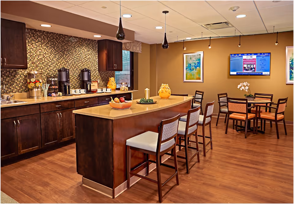 A cozy dining and kitchen area with a central island featuring four chairs. The kitchen has dark wooden cabinets, a mosaic tile backsplash, and a countertop with coffee dispensers, cups, and a water dispenser with lemon slices. The room has wooden flooring, warm beige walls, framed artwork, and a wall-mounted TV displaying information.