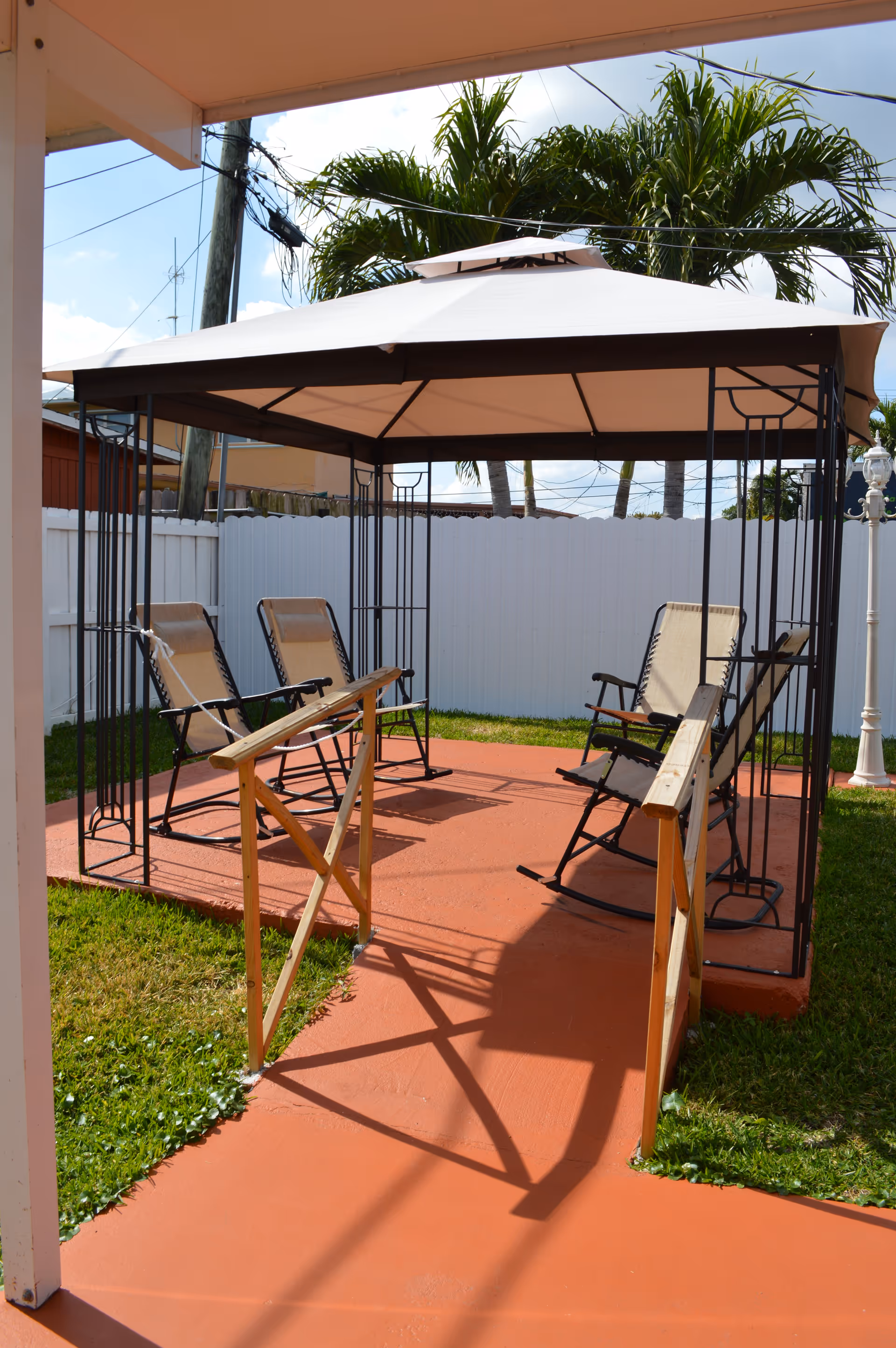 Outdoor patio area with a gazebo featuring a beige canopy and four reclining chairs arranged underneath. The area has a red concrete floor with wooden handrails leading into the gazebo. There is a white fence and palm trees in the background under a partly cloudy sky.