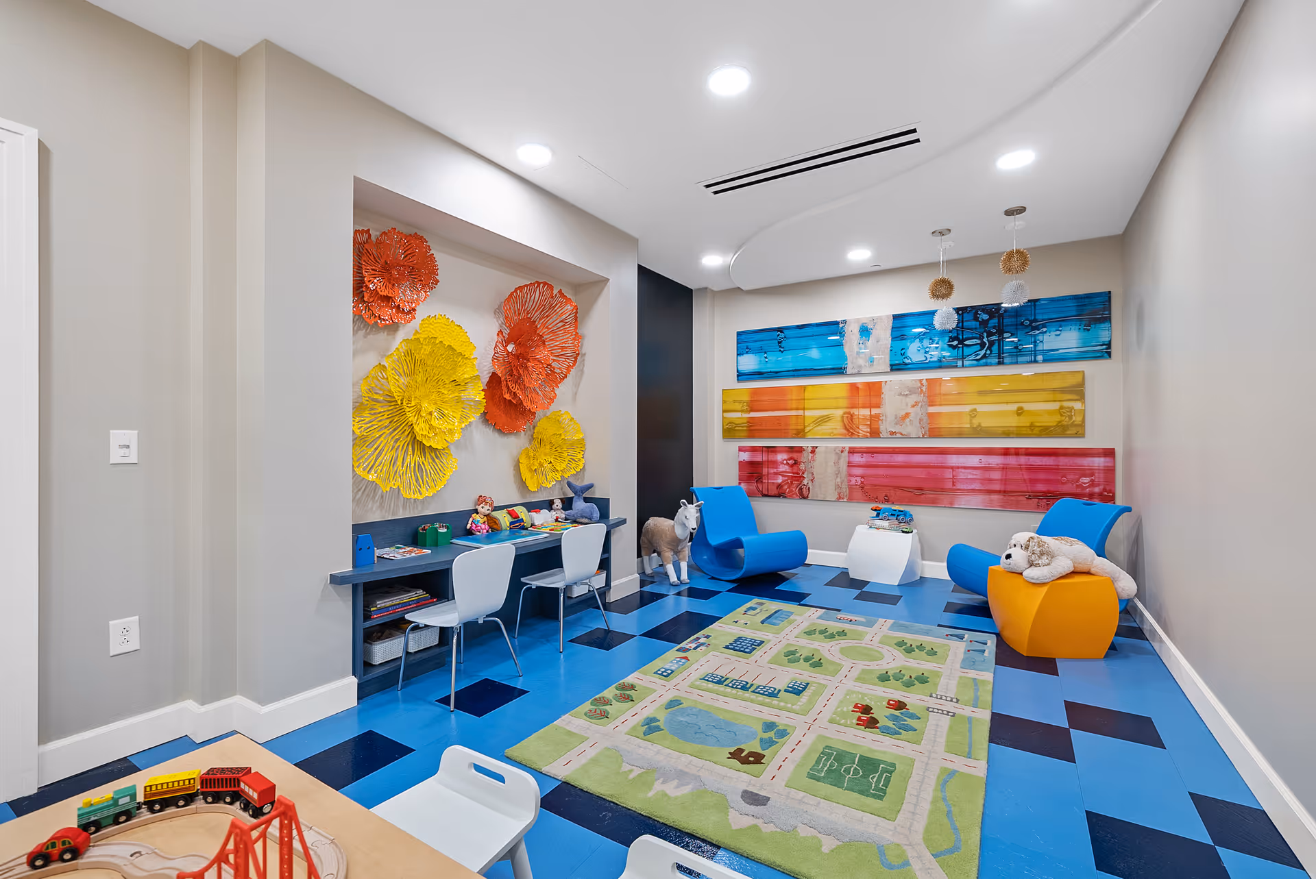 A colorful children's playroom with blue and black checkered flooring, a green play rug featuring roads and buildings, blue and yellow modern chairs, a small white table, and a wall-mounted desk with two white chairs. The walls are decorated with large, vibrant yellow and orange flower sculptures and three horizontal abstract art panels in blue, yellow, and red. Toys including stuffed animals and a wooden train set are visible.