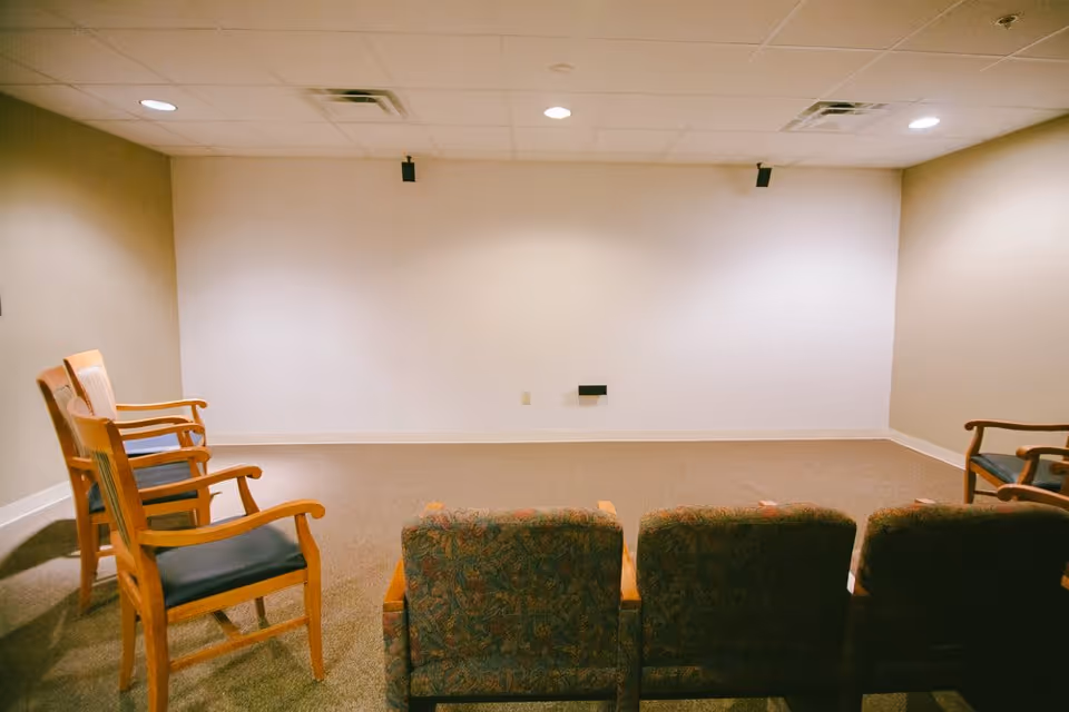 A small room with a beige carpet and plain white walls, furnished with two rows of chairs facing a blank wall. The front row has three cushioned chairs with patterned fabric, and the back row has four wooden chairs with blue cushions. The ceiling has recessed lighting and air vents.