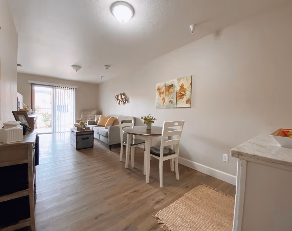 A bright living area with wooden flooring featuring a small round dining table with two white chairs, a gray sofa with beige cushions, a decorative wall art, and a sliding glass door leading outside. The room has neutral-colored walls and ceiling lights.