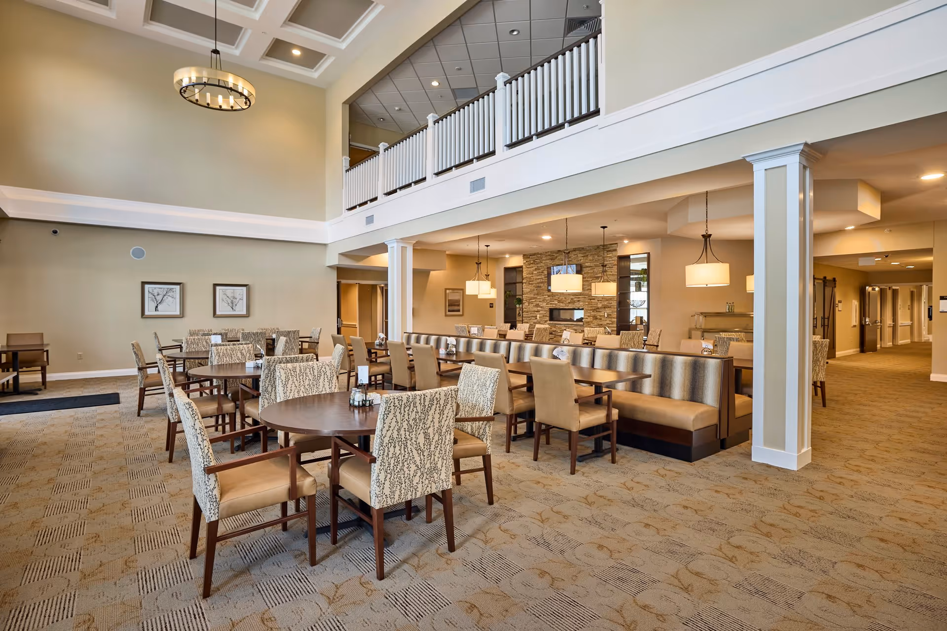 Spacious dining room with multiple round and rectangular tables surrounded by cushioned chairs. The room features a high ceiling with a modern chandelier, beige walls, carpeted floor, and a stone accent wall with a fireplace and pendant lights. There is a balcony railing visible on the upper level and a hallway leading to other areas.