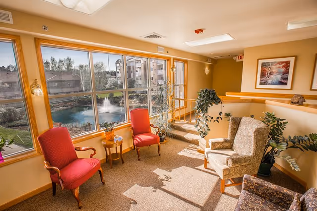 A bright and cozy sitting area in a senior living facility with large windows overlooking a pond with a fountain and surrounding buildings. The room features two red upholstered armchairs, a small wooden side table with a pineapple decoration, a patterned armchair, and several potted plants. The walls are painted a warm yellow and decorated with framed artwork.
