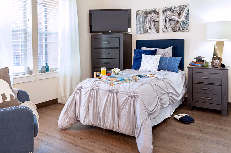 Bright, tastefully decorated bedroom with a single bed with light bedding and blue pillows, a breakfast tray on the bed, dressers, a TV, and a window with curtains.