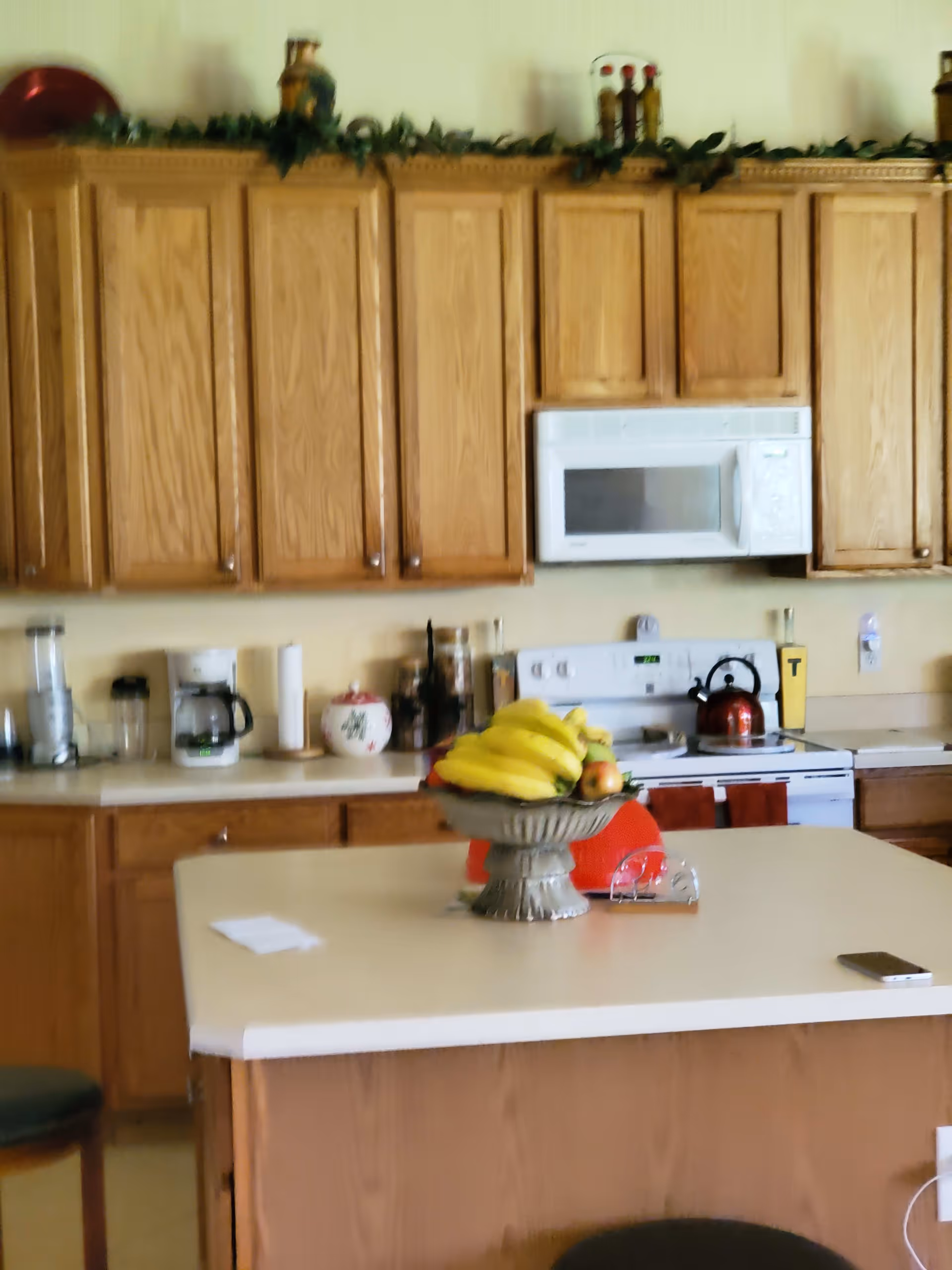 A kitchen with wooden cabinets, a white microwave above a white stove, and a countertop island in the center. On the island, there is a decorative bowl filled with bananas and other fruit. Various kitchen appliances and items are on the counters, including a coffee maker, blender, and a red kettle on the stove.