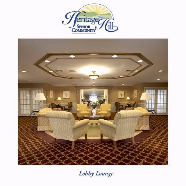 A spacious and well-lit lobby lounge area with patterned carpet flooring, beige upholstered armchairs and sofas arranged around a central coffee table with a flower vase. The ceiling features a decorative recessed design with a central light fixture. The room has multiple lamps, framed artwork on the walls, and glass-paneled double doors on either side.