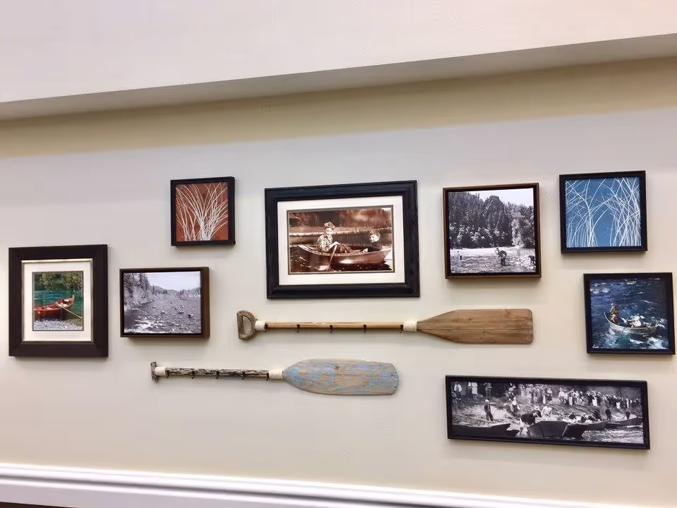 A wall decorated with a collection of framed pictures and two wooden oars. The pictures include black and white and color images of people in boats, nature scenes, and abstract designs. The frames are arranged in a rectangular pattern on a light-colored wall.