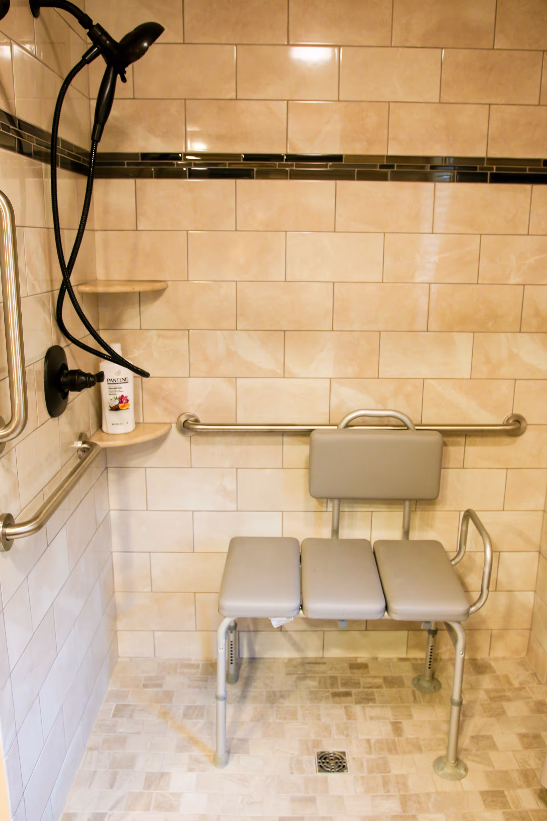 Accessible shower area with beige tiled walls and floor, equipped with a foldable shower bench with padded seat and backrest, grab bars on the walls, a handheld showerhead, and a small corner shelf holding a bottle of Pantene shampoo.
