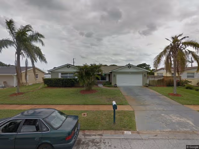 Single-story residential house with a two-car garage, a driveway, and a front yard featuring palm trees and trimmed bushes under a cloudy sky. A green car is parked on the street in front of the house.