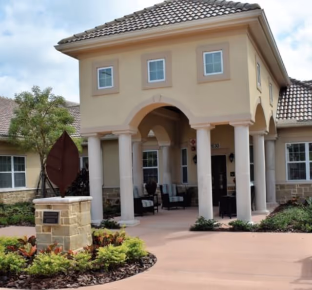 Entrance of a senior living facility with a covered porch supported by columns, outdoor seating, and landscaped garden beds with a decorative leaf sculpture on a stone pedestal.