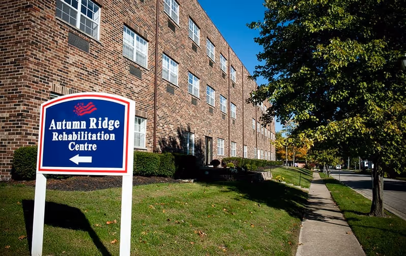 Exterior view of a brick building with multiple windows, a sidewalk, and a green lawn with bushes and trees. A blue and white sign in the foreground reads 'Autumn Ridge Rehabilitation Centre' with an arrow pointing left.