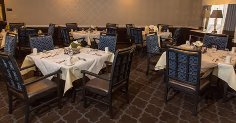 A dining room with multiple tables covered in white tablecloths, each set with plates, cups, and silverware. The chairs have dark wooden frames with patterned blue upholstery. The room has a carpeted floor with a geometric pattern and neutral-colored walls with subtle wallpaper. A window with blinds and a lamp on a side table are visible in the background.