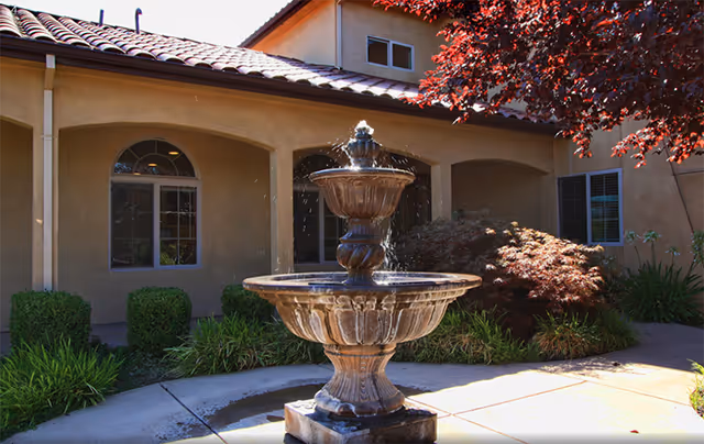 Stone three-tier fountain in a landscaped courtyard in front of a stucco building with arched walkways and a tiled roof.