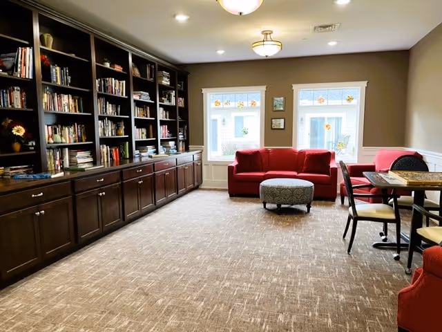 Common lounge with built-in bookshelves, red sofas and ottoman, and a small table with chairs by two windows.