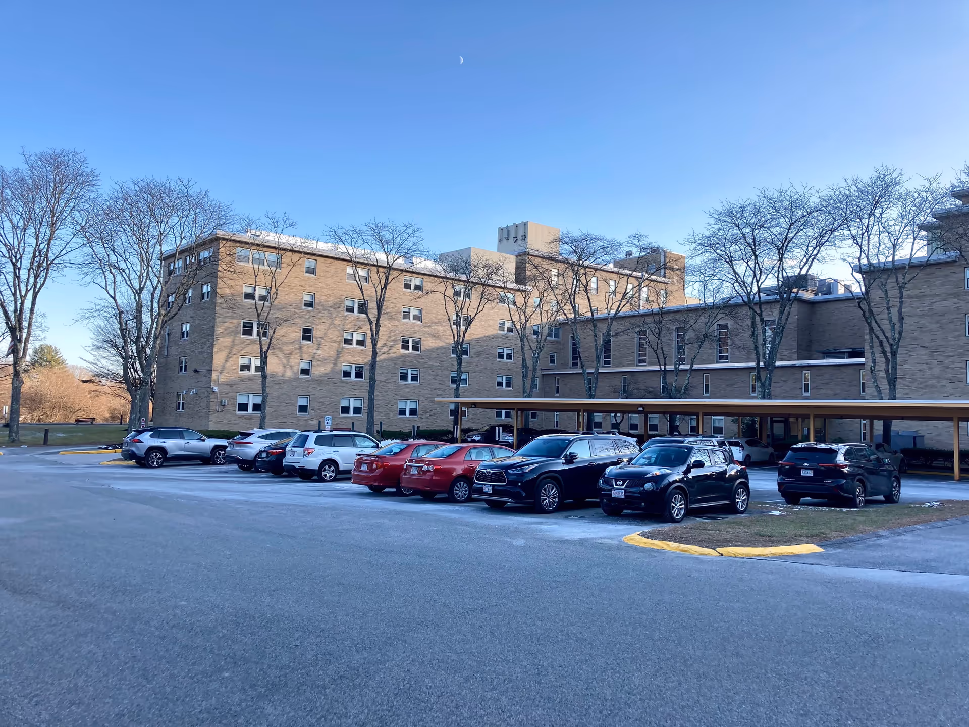 Exterior view of a multi-story brick building with several windows, surrounded by leafless trees and a parking lot with multiple parked cars under a clear blue sky.