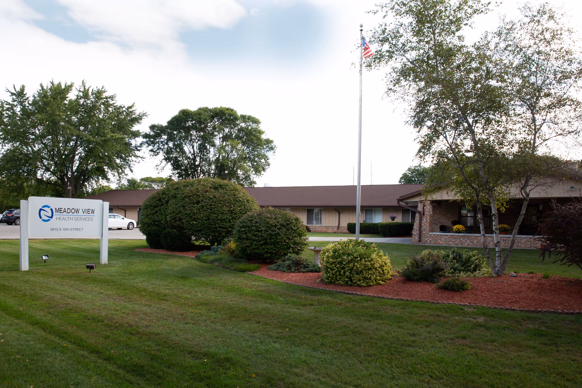 Exterior view of Meadow View Health Services building with a well-maintained lawn, bushes, trees, an American flag on a flagpole, and a sign displaying the facility's name and address.