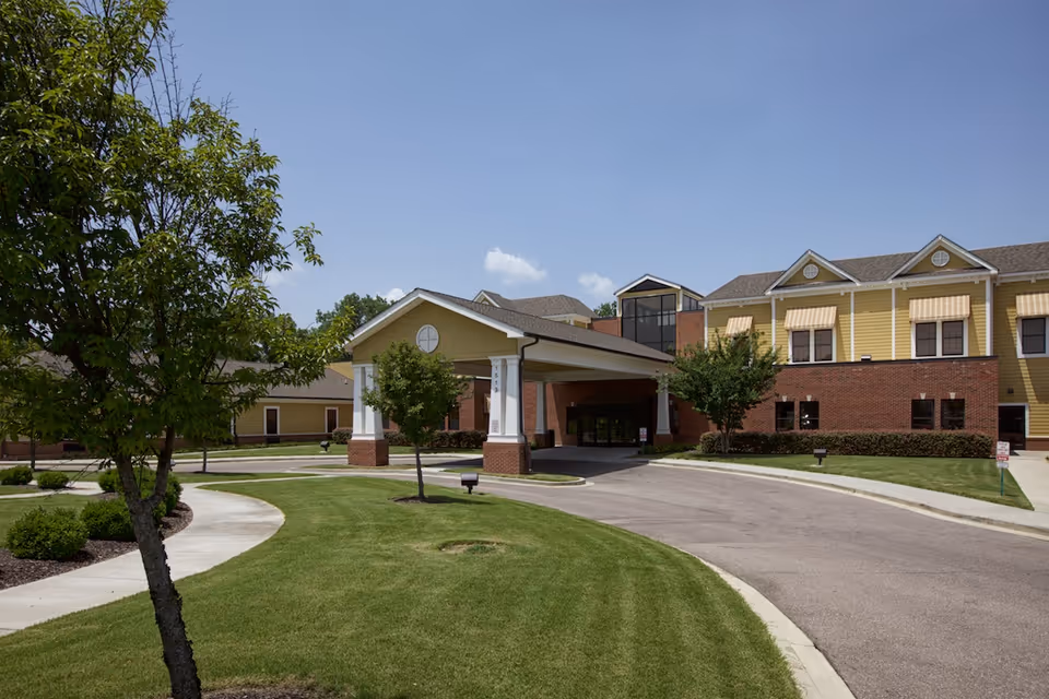 Exterior view of AHC Harbor View facility showing a driveway leading to a covered entrance with a yellow and brick building in the background, surrounded by green grass, trees, and a clear blue sky.
