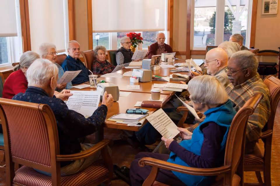A group of elderly people sitting around a large wooden table in a well-lit room, each holding and reading from sheets of paper, possibly participating in a group activity or meeting.