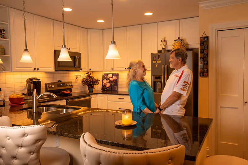 An elderly couple holding hands and smiling at each other in a modern kitchen with white cabinets, granite countertops, pendant lights, and stainless steel appliances.