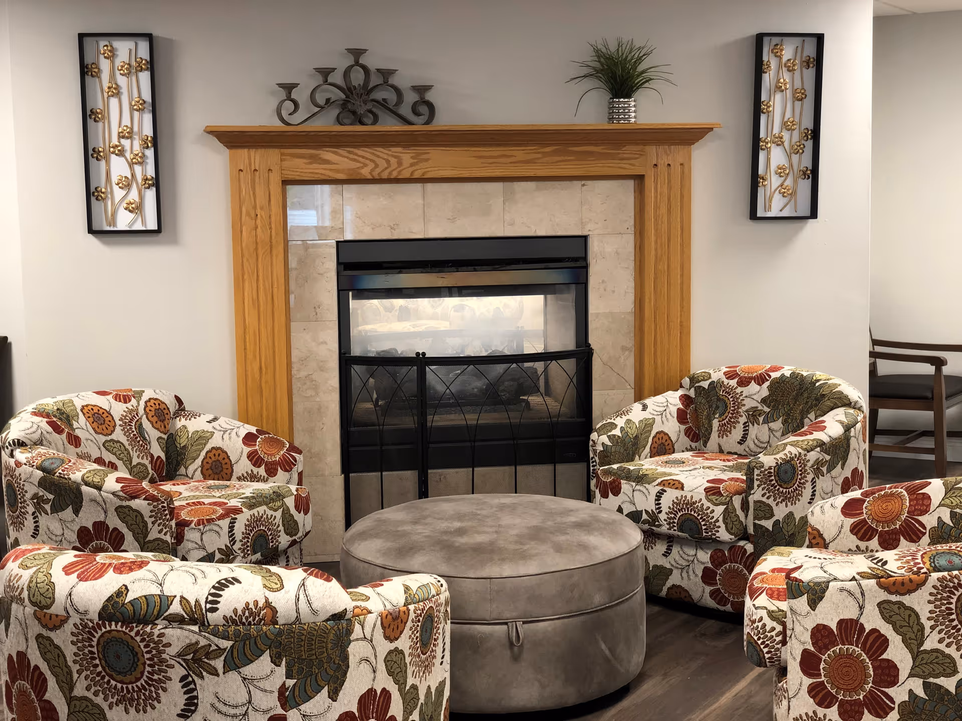 A cozy seating area with four floral-patterned armchairs arranged around a round gray ottoman in front of a fireplace with a wooden mantel. The fireplace has a black metal screen and is surrounded by beige tiles. On the mantel, there is a small potted plant and a decorative metal candle holder. Two framed wall decorations with gold floral designs hang on either side of the fireplace.