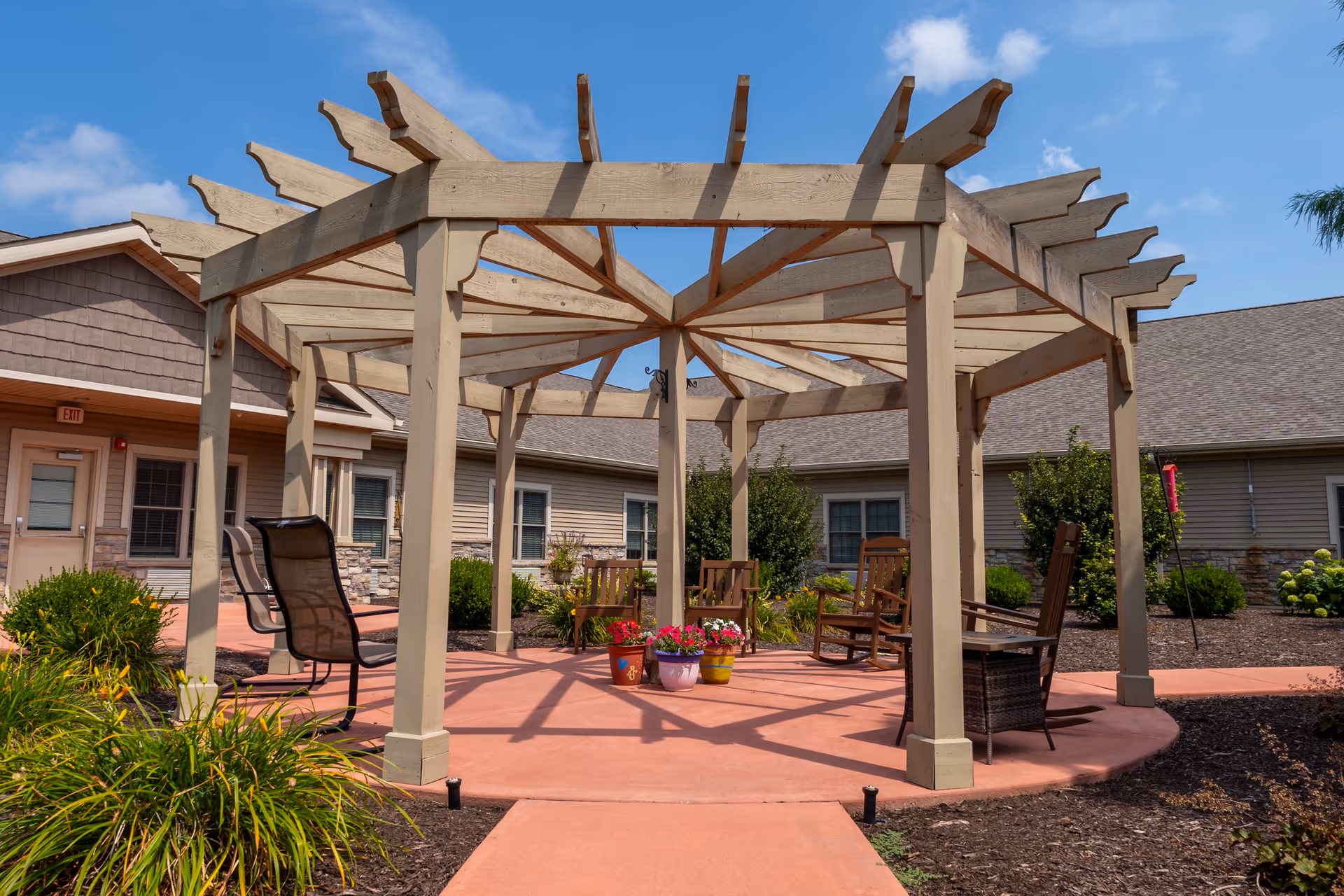 Outdoor seating area with a wooden pergola structure surrounded by a circular concrete patio. Several chairs, including rocking chairs and a mesh chair, are arranged under the pergola. Potted flowers are placed in the center of the patio. The area is landscaped with bushes and plants, and single-story buildings with beige siding and stone accents are visible in the background under a partly cloudy blue sky.