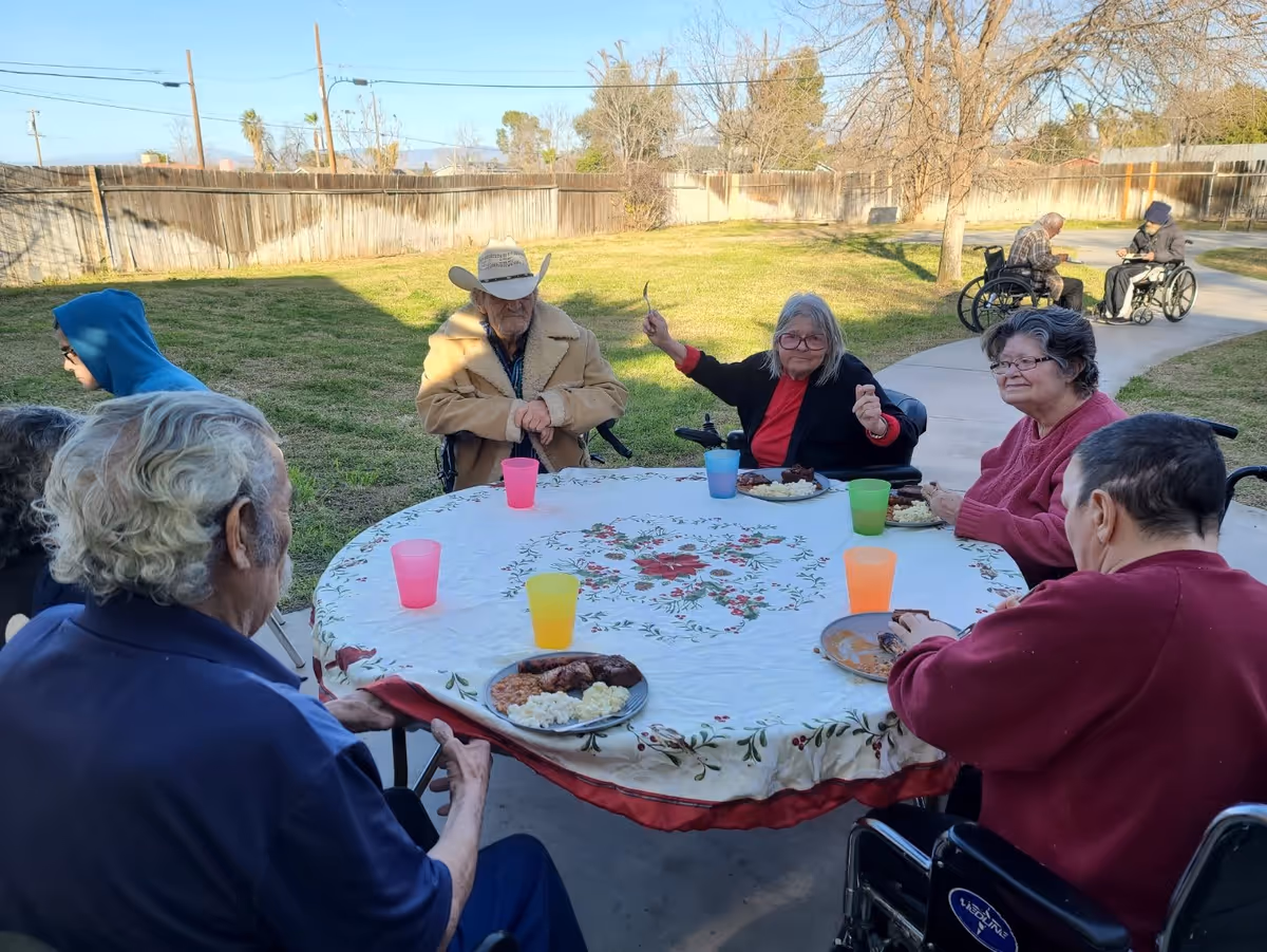 A group of elderly people sitting around a round table outdoors, enjoying a meal together. The table is covered with a floral tablecloth and has colorful plastic cups. Some individuals are in wheelchairs, and the setting is a grassy yard with a wooden fence and a paved walkway in the background.