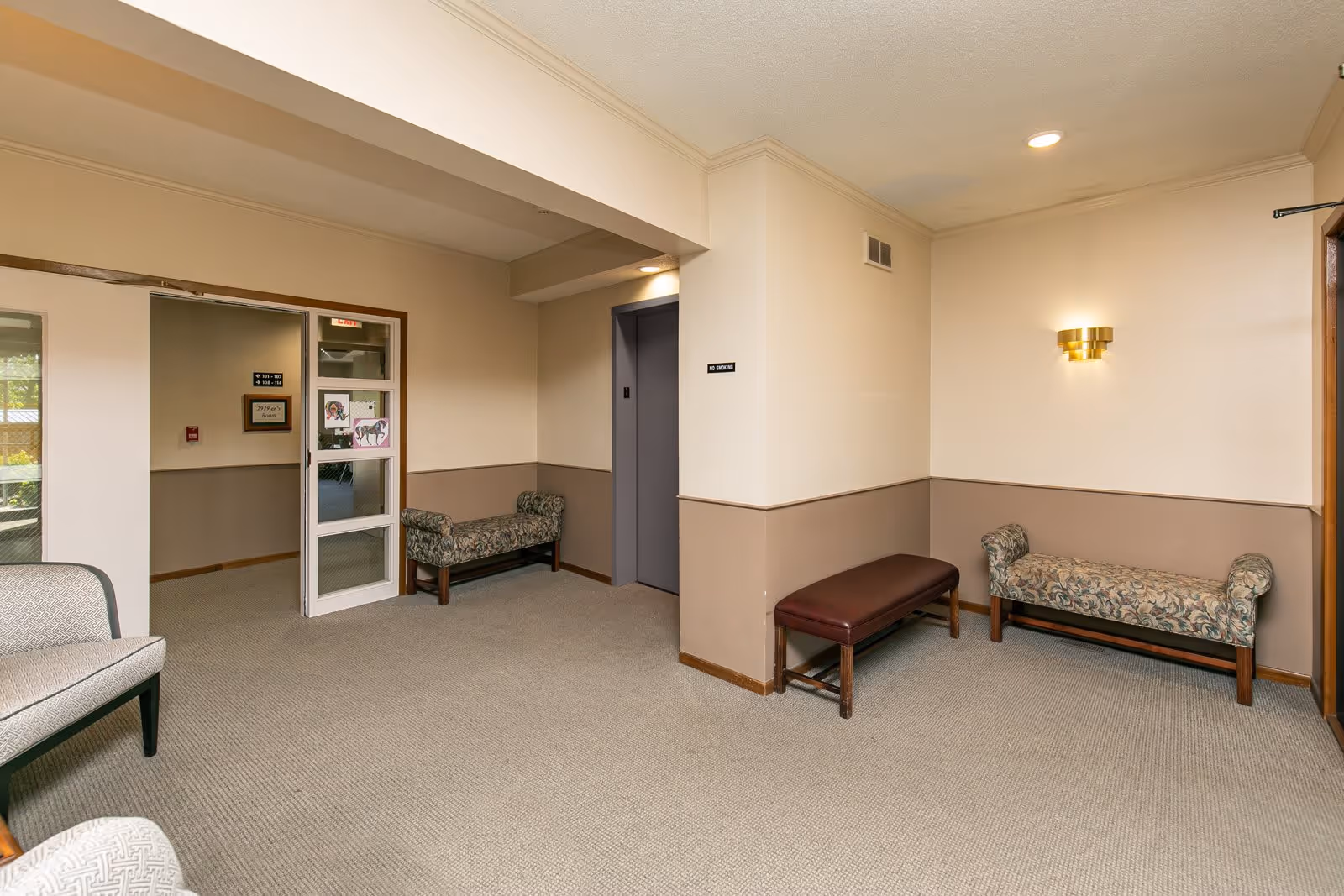 Interior view of a senior apartment building hallway with beige walls and carpeted floor. There are several benches with patterned upholstery and a brown leather bench. A gray elevator door is visible with a 'No Smoking' sign above it. A doorway with glass panels leads to another hallway with signs indicating room numbers. A gold wall sconce light fixture is mounted on the wall.