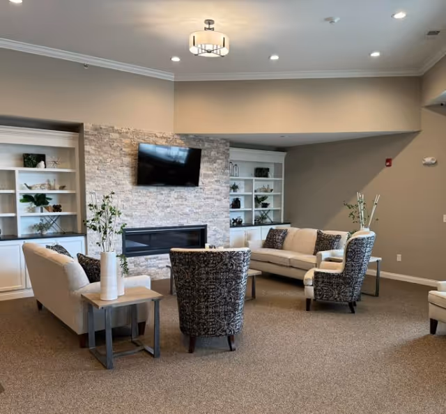 Spacious assisted-living common room with couches and chairs arranged around a wall-mounted TV above a fireplace and built-in shelving.