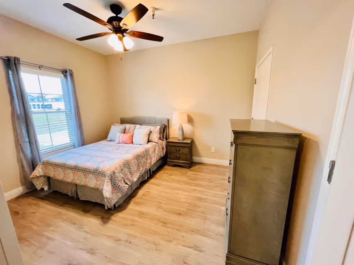 A bedroom with a bed covered in a patterned quilt and several pillows, a nightstand with a lamp, a wooden dresser, a ceiling fan with lights, and a window with curtains letting in natural light.