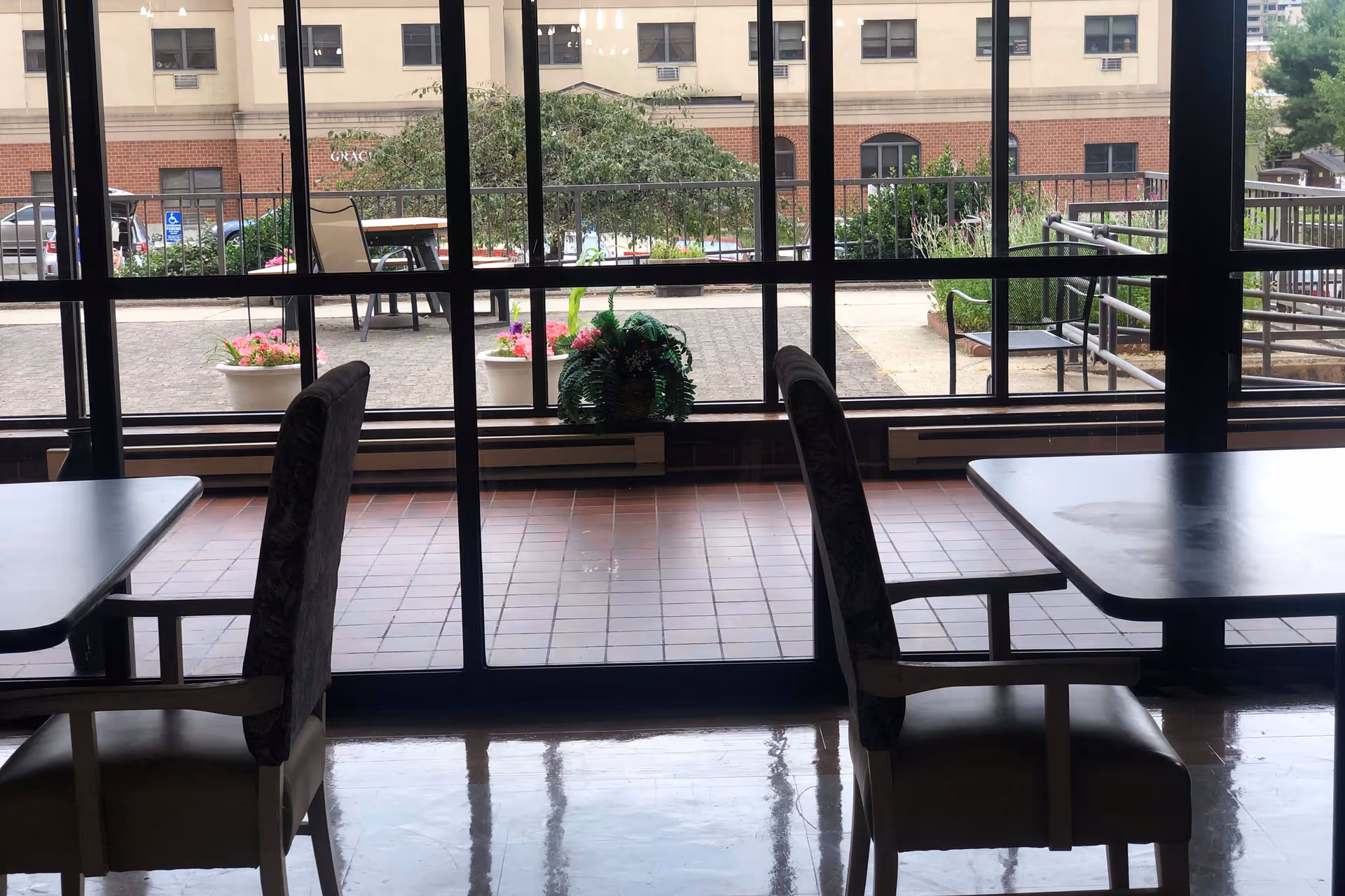 View from inside a room with tables and chairs looking out through large glass windows onto an outdoor patio area with potted plants and outdoor seating. A building is visible in the background.