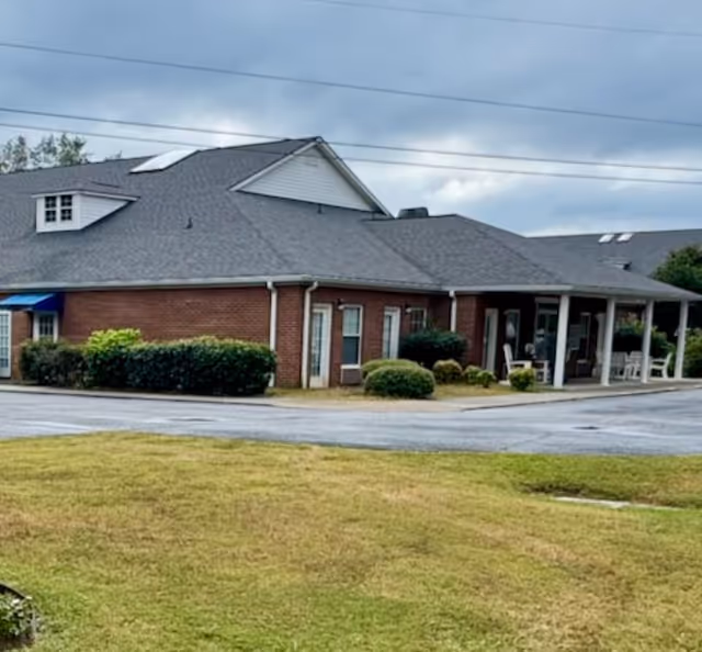 Exterior view of a single-story brick building with a gray roof, surrounded by grass and bushes under a cloudy sky. The building has a covered porch area with chairs and tables.