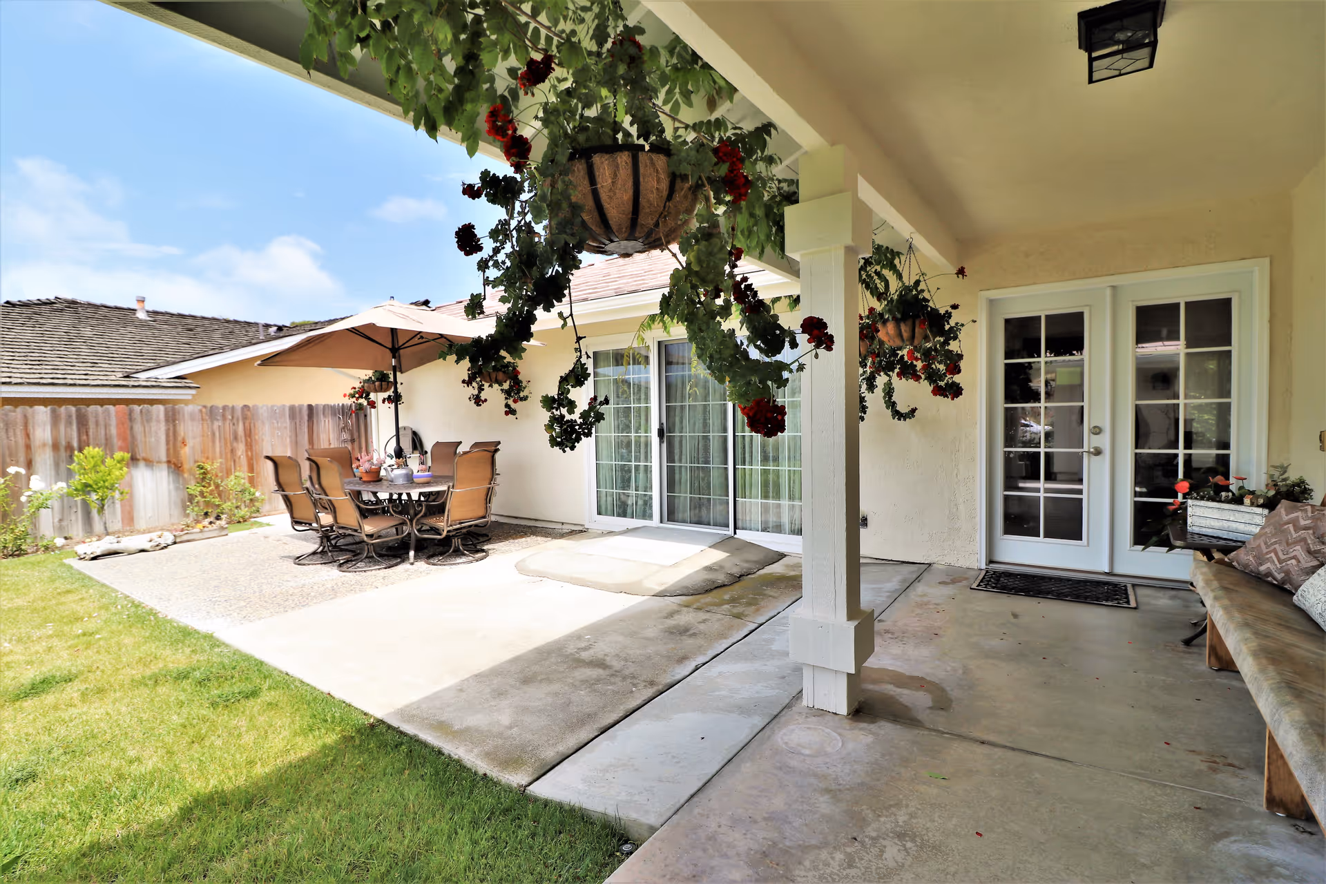Covered patio area with hanging flower baskets, a wooden bench with cushions, and a concrete floor. Adjacent to the patio is an outdoor dining area with a table, six chairs, and a large umbrella. The area is enclosed by a wooden fence and has a grassy lawn under a partly cloudy sky.