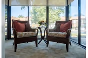 Two wicker armchairs with cushions flank a small table in a sunlit seating area with floor-to-ceiling windows showing trees outside.