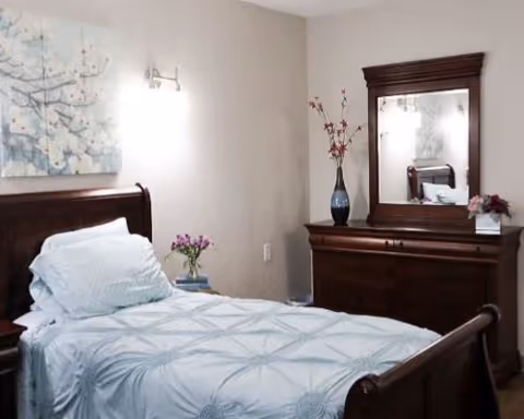 A cozy bedroom with a wooden bed frame and a neatly made bed with white and light blue bedding. There is a wooden dresser with a mirror on top, decorated with a vase of flowers and a small floral arrangement. A soft wall light is mounted above the bed, and a floral painting hangs on the wall.