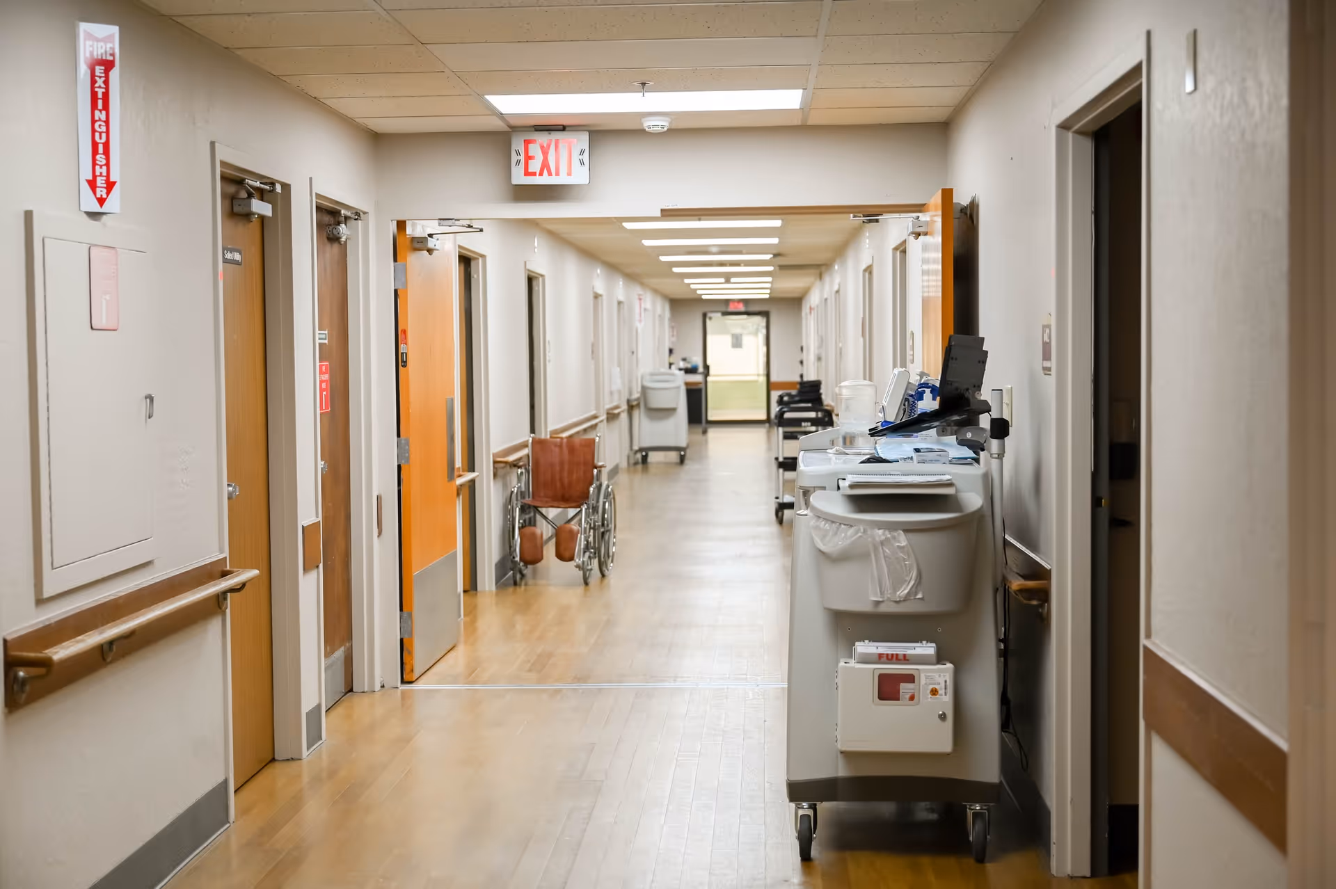 A long, well-lit hallway in a care facility with wooden floors and beige walls. Several closed doors line both sides of the corridor. A wheelchair is positioned near an open door on the left side. Medical carts and equipment are placed along the right side of the hallway. An exit sign is visible on the ceiling near the center of the image.