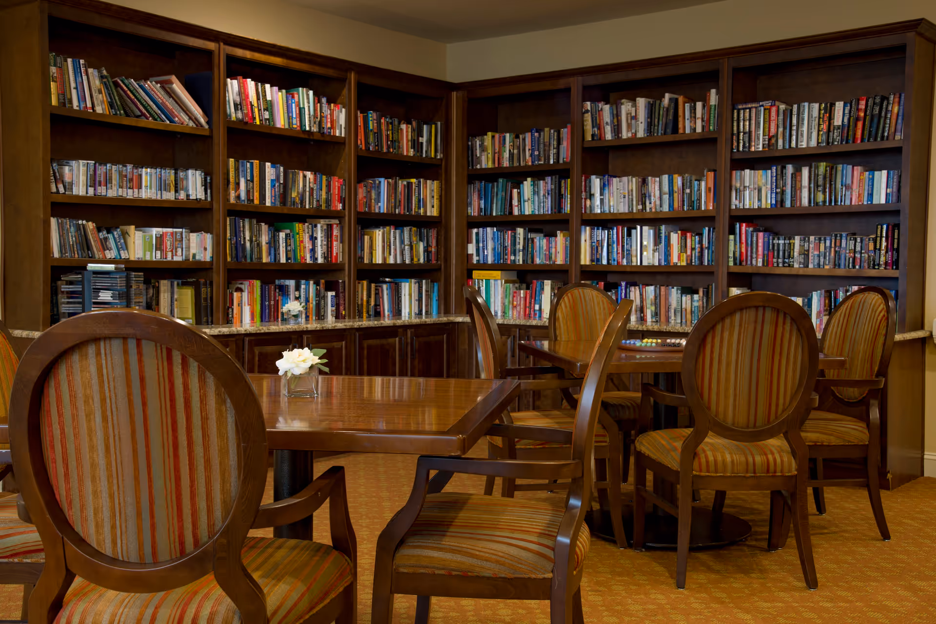 A cozy library room with wooden bookshelves filled with books lining the walls. Several wooden tables with striped upholstered chairs are arranged in the room, and a small vase with white flowers is placed on one of the tables.